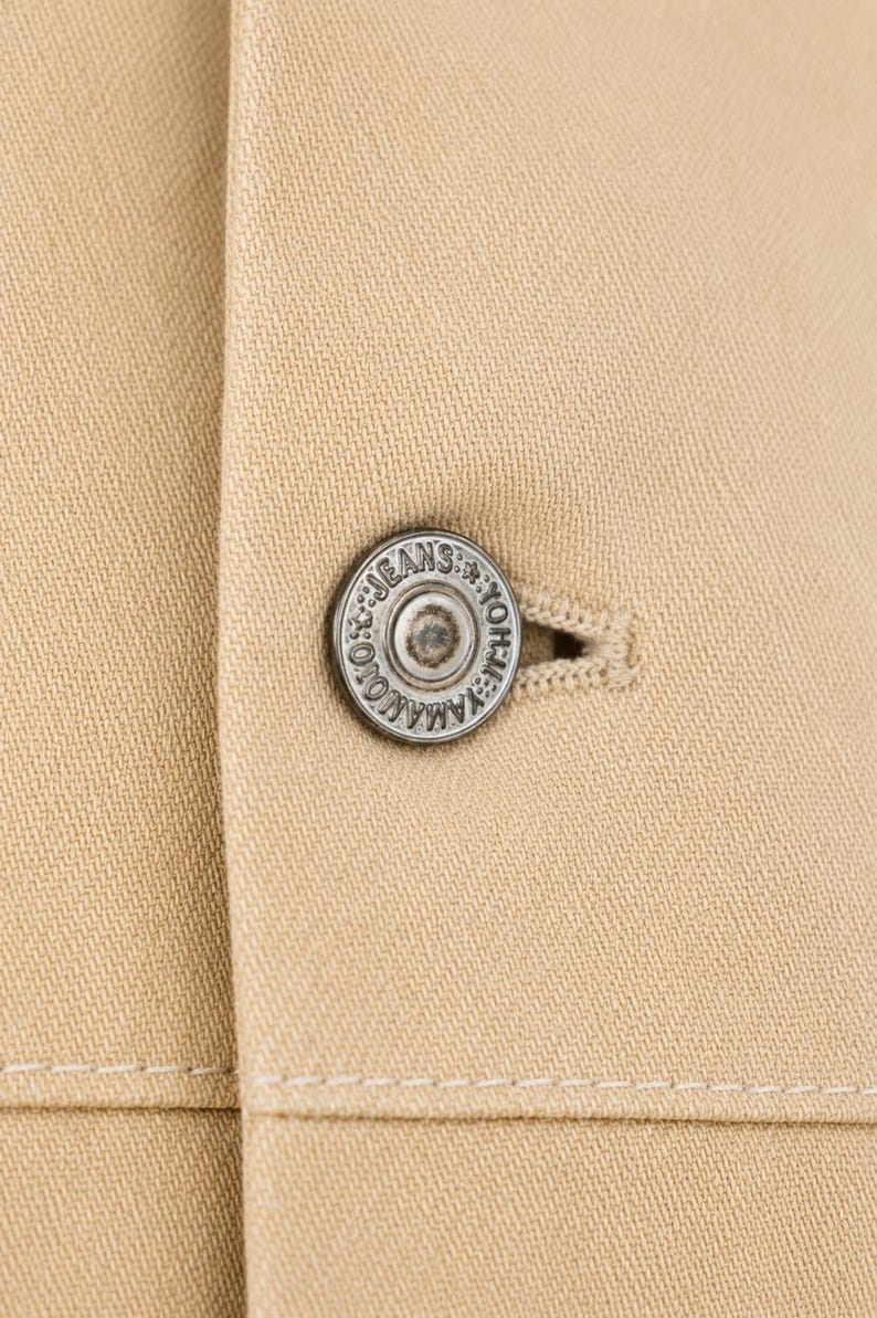 May include: Close-up of a beige jacket featuring a silver button. The button is circular and has the text "YAMAMOTO JEANS" around the edge. The jacket's fabric is a plain beige with a visible weave.