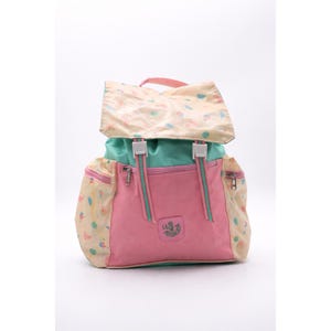 May include: A colorful backpack with a flap closure. The bag features a pink front, a teal center section, and a flap with a light yellow background and a pattern of small, colorful shapes. The bag has two side pockets and adjustable straps.