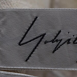 May include: A close-up shot of a grey fabric label with black embroidered text. The label is attached to a beige woven fabric. The text appears to be a signature or brand name.