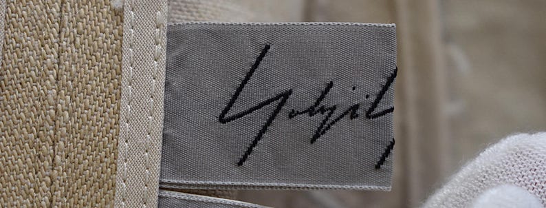 May include: A close-up shot of a grey fabric label with black embroidered text. The label is attached to a beige woven fabric. The text appears to be a signature or brand name.