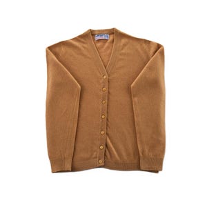 Puede incluir: Un cárdigan de color beige con cuello en V y cierre de botones en la parte delantera. El suéter de manga larga tiene puños y dobladillo acanalados. El suéter está hecho de un material suave y está plano sobre una superficie blanca.