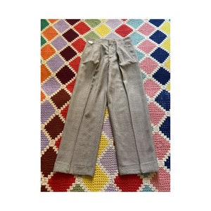 Puede incluir: Pantalones de cuadros grises claros con diseño de pernera ancha. Los pantalones tienen pliegues en la cintura y cierre de botón. La tela tiene un sutil estampado de cuadros. Los pantalones se muestran sobre una colorida manta estampada.