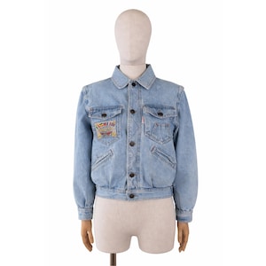 Può includere: Giacca di jeans azzurra con chiusura a bottoni, due tasche sul petto e due tasche laterali. La giacca presenta una piccola toppa ricamata sul petto sinistro e un colletto rotondo. La giacca è esposta su un manichino.
