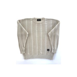 Puede incluir: Un suéter de punto acanalado beige con cuello redondo. El suéter tiene un patrón de rayas verticales y mangas largas. La marca "VERSACE" es visible en una pequeña etiqueta en el cuello y otra etiqueta en la parte inferior izquierda.