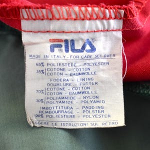 May include: A close-up of a Fila clothing tag. The tag is white with blue text and the Fila logo. The tag reads "Made in Italy" and lists the fabric composition in multiple languages. The tag is against a red and green fabric.