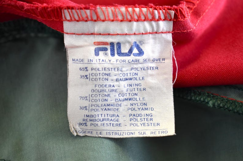 May include: A close-up of a Fila clothing tag. The tag is white with blue text and the Fila logo. The tag reads "Made in Italy" and lists the fabric composition in multiple languages. The tag is against a red and green fabric.