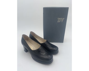 May include: Pair of black leather platform shoes with a square toe and a chunky heel. The shoes are sitting on a gray surface next to a box with the letters "DV" on it.