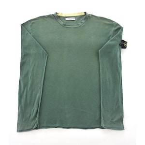 May include: A long-sleeved, olive green shirt with a crew neck and a small logo patch on the left sleeve. The shirt has a relaxed fit and a faded appearance. The collar has a yellow lining. The brand name "STONE ISLAND" is printed on the collar.