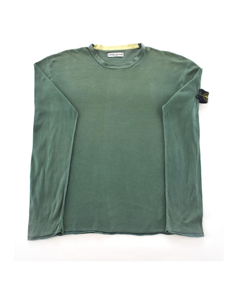 May include: A long-sleeved, olive green shirt with a crew neck and a small logo patch on the left sleeve. The shirt has a relaxed fit and a faded appearance. The collar has a yellow lining. The brand name "STONE ISLAND" is printed on the collar.