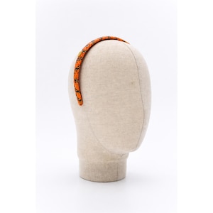 Może przedstawiać: Opaska do włosów we wzór pomarańczowo-czarny jest pokazana na głowie manekina. Opaska ma powtarzający się wzór czarnych kształtów na pomarańczowym tle. Głowa manekina ma neutralny beżowy kolor.