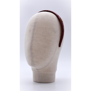 Może przedstawiać: Bordowa pleciona opaska do włosów z małym, złotym emblematem. Opaska jest prezentowana na głowie manekina. Akcesorium jest przeznaczone do noszenia na głowie.