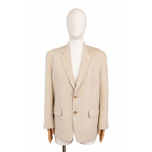 Può includere: Un blazer beige con chiusura a due bottoni, revers a lancia e due tasche anteriori. La giacca è esposta su un manichino con braccia in legno. Il blazer è realizzato in un tessuto colorato chiaro.