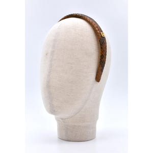 Może przedstawiać: Brązowa opaska do włosów ze skóry węża ze złotym emblematem. Opaska jest prezentowana na głowie manekina na białym tle. Wzór skóry węża ma odcienie brązu i czerni.