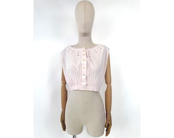May include: A light pink sleeveless crop top with a button-up front. The top features delicate eyelet detailing around the neckline and vertical pleats. The garment is displayed on a mannequin.