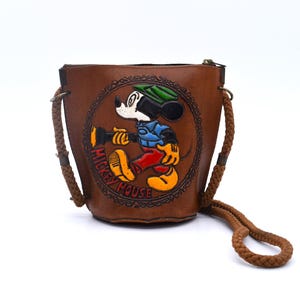 Puede incluir: Bolso de cubo de cuero marr&oacute;n con una imagen en relieve de Mickey Mouse. El bolso tiene una correa trenzada para el hombro y un cierre de cremallera. La imagen de Mickey Mouse est&aacute; rodeada por las palabras "Mickey Mouse" en un dise&ntilde;o circular.