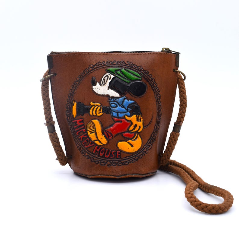 Puede incluir: Bolso de cubo de cuero marr&oacute;n con una imagen en relieve de Mickey Mouse. El bolso tiene una correa trenzada para el hombro y un cierre de cremallera. La imagen de Mickey Mouse est&aacute; rodeada por las palabras "Mickey Mouse" en un dise&ntilde;o circular.