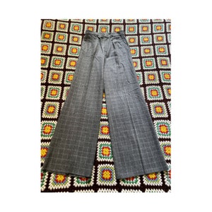 Può includere: Pantaloni grigi a gamba larga con motivo a quadri. I pantaloni hanno una vita alta e un classico motivo a quadri. Il tessuto sembra essere un materiale di peso medio, adatto alle temperature più fresche. I pantaloni sono esposti su una superficie colorata e fantasia.