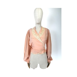 Puede incluir: Una blusa de color melocotón con mangas largas y un ribete de encaje blanco alrededor del cuello y el dobladillo.