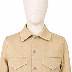 May include: A beige denim jacket with two chest pockets and silver buttons. The jacket is displayed on a mannequin. The jacket has a classic collar and a button-up front. The fabric appears to be a medium weight denim.