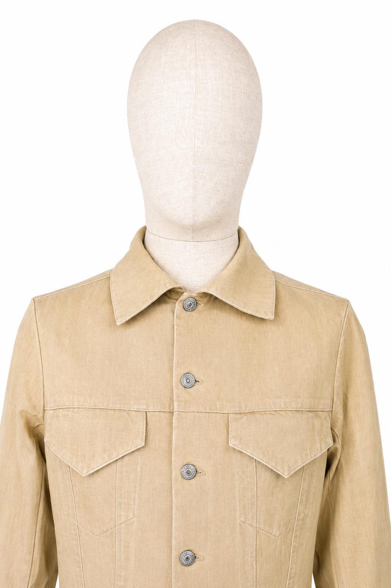 May include: A beige denim jacket with two chest pockets and silver buttons. The jacket is displayed on a mannequin. The jacket has a classic collar and a button-up front. The fabric appears to be a medium weight denim.