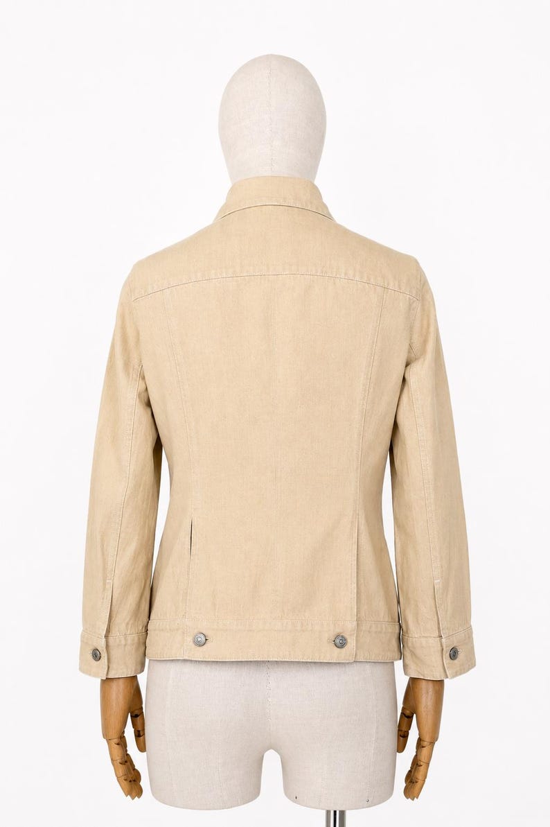 May include: A beige jacket with a button-down front, long sleeves, and two small pockets. The jacket has a classic design with a collar and buttoned cuffs. The jacket is displayed on a mannequin.