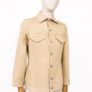 May include: A beige denim jacket with a traditional collar, two chest pockets, and button fastenings. The jacket is displayed on a mannequin and has long sleeves. The shorts are also beige. Measurements are not visible.