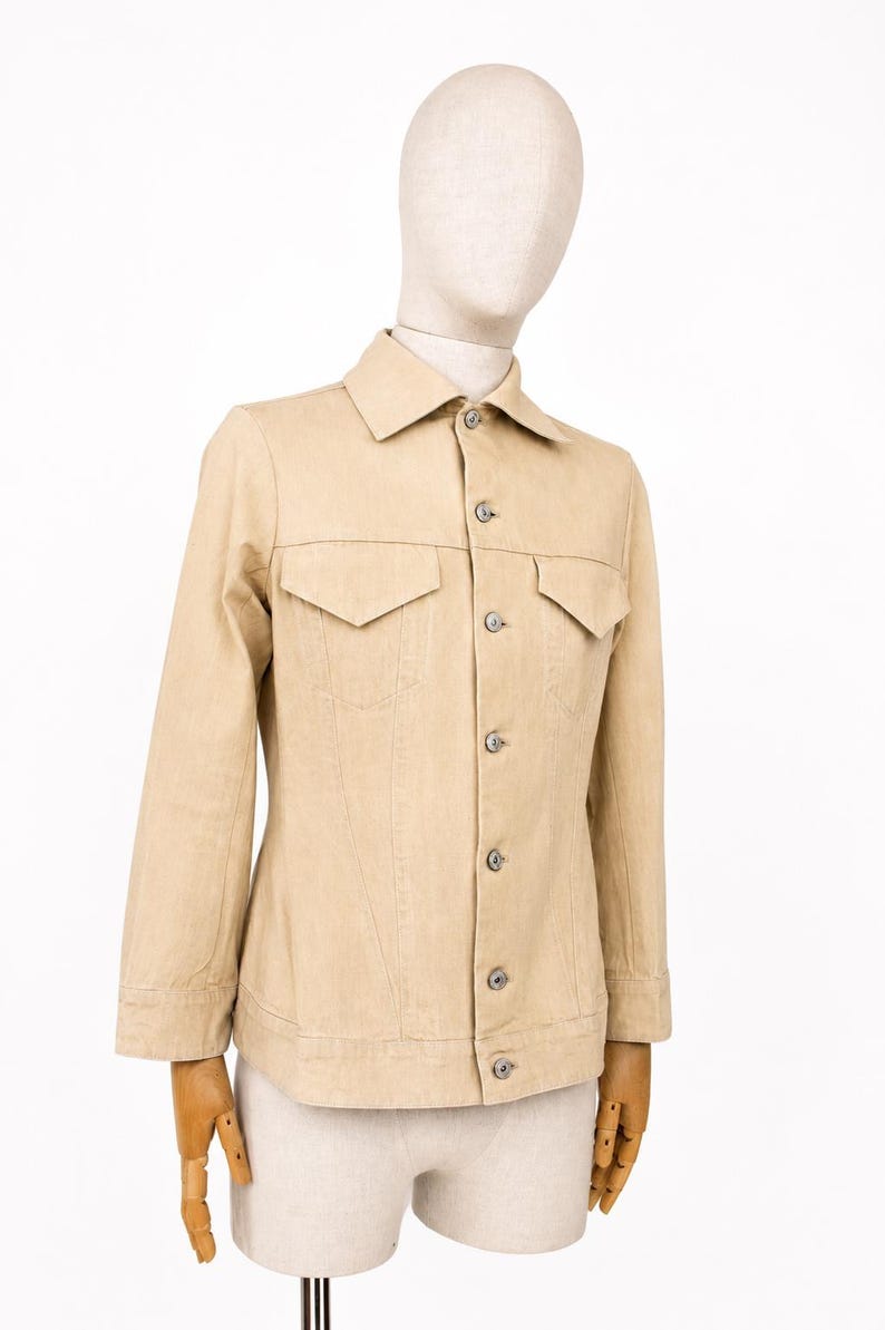 May include: A beige denim jacket with a traditional collar, two chest pockets, and button fastenings. The jacket is displayed on a mannequin and has long sleeves. The shorts are also beige. Measurements are not visible.