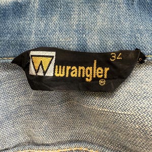 以下が含まれることがあります： 白い三角形と金色の文字で「Wrangler」と書かれた黒と金の Wrangler ブランドラベル。ラベルには、白い文字で「34」と書かれたサイズタグも付いています。