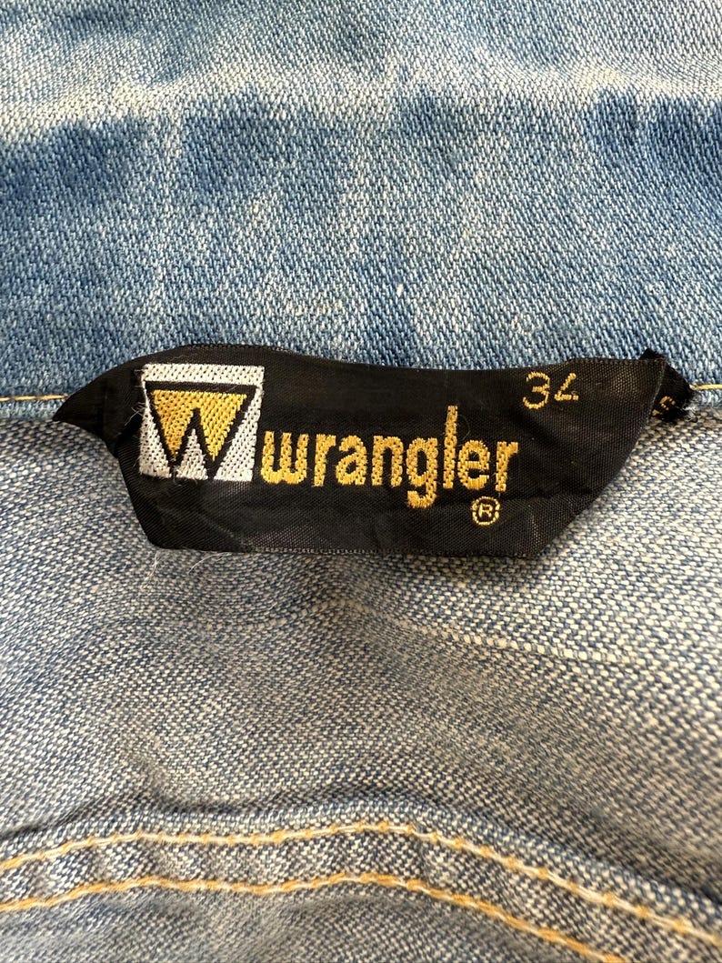 以下が含まれることがあります： 白い三角形と金色の文字で「Wrangler」と書かれた黒と金の Wrangler ブランドラベル。ラベルには、白い文字で「34」と書かれたサイズタグも付いています。