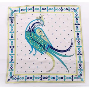 May include: Square white scarf with a peacock design. The peacock is in shades of blue and teal, with a yellow crown and tail. The scarf has a border with blue and yellow geometric shapes and small blue dots.