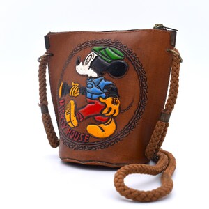 Puede incluir: Bolso tipo cubo de cuero marr&oacute;n con una imagen en relieve de Mickey Mouse. El bolso tiene una correa de hombro de cuerda y cierre de cremallera. Las palabras "Mickey House" est&aacute;n impresas.