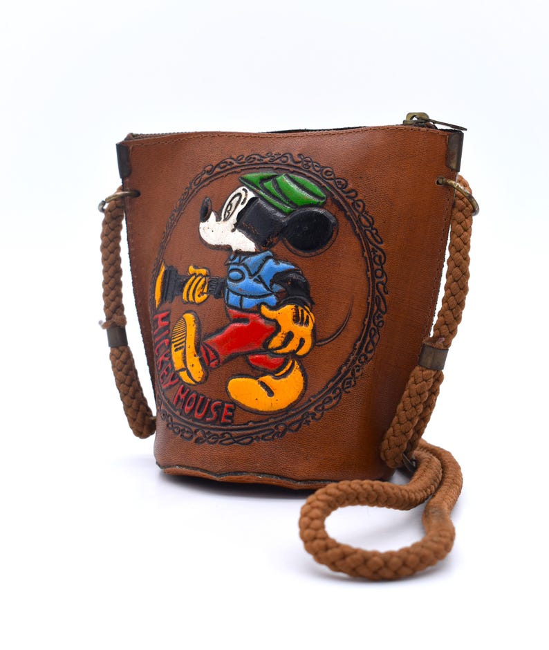 Puede incluir: Bolso tipo cubo de cuero marr&oacute;n con una imagen en relieve de Mickey Mouse. El bolso tiene una correa de hombro de cuerda y cierre de cremallera. Las palabras "Mickey House" est&aacute;n impresas.