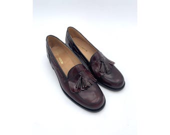Pode incluir: Um par de mocassins de couro bordô com detalhes de borlas. Os sapatos têm um design clássico com bico redondo e salto baixo. Os mocassins são exibidos em um fundo branco.
