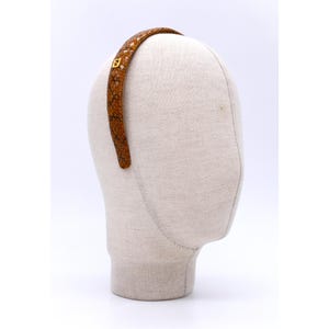 Może przedstawiać: Brązowa opaska do włosów ze wzorem skóry węża i złotym kwadratowym akcentem. Zakrzywiona opaska jest prezentowana na głowie manekina. Wzór skóry węża zawiera odcienie brązu i czerni.