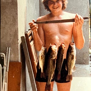 May include: A young person wearing a blue baseball cap holds a wooden stick and five fish. The fish are hanging from the stick and are all about the same size. The person is wearing black shorts and is standing in front of a wooden door.