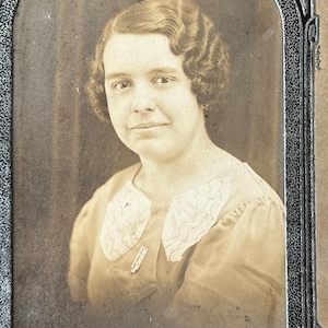 May include: A sepia-toned portrait of a woman with wavy hair, wearing a light-colored blouse with a lace collar. The photo is framed in a black and gold frame.