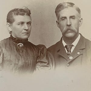 May include: Sepia-toned portrait of a man and woman. The woman wears a dark dress with a high collar and a brooch. The man has a large mustache, a suit jacket, and a tie. The photograph appears to be from the late 19th or early 20th century.