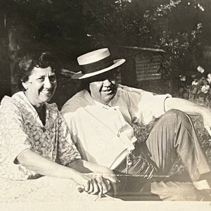 May include: A black and white photo of a couple sitting outdoors. The woman is wearing a patterned dress and the man is wearing a white shirt, a straw hat, and pants.
