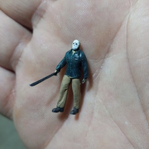 May include: Miniature figure of a horror movie character, wearing a white mask, dark jacket, tan pants, and black boots. The figure is holding a black machete. The figure is approximately 1.5 inches tall.