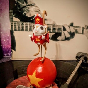 May include: A red and white figurine of a mouse dressed as a drummer, balancing on a red ball with a yellow star. The figurine is on a black vinyl record with a record player needle.