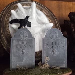 Puede incluir: Dos decoraciones grises en forma de lápida con los nombres "Eric Draven" y "Shelly Webster" inscritos en ellas. Una figurita de cuervo negro se sienta sobre una escultura de mano blanca detrás de las lápidas.