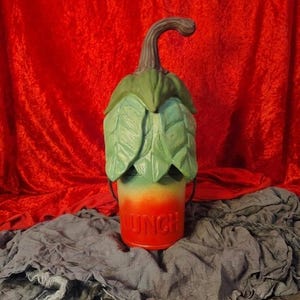 May include: A cylindrical lunch pail featuring a lid shaped like a green leafy vegetable top with a curved brown stem. The base of the container transitions from a light green to a vibrant orange color, with the word LUNCH embossed in capital letters.