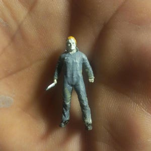 May include: A small, detailed figurine of a horror movie character wearing a blue jumpsuit and a white mask. The figurine is holding a knife in one hand.