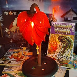 May include: A red glass leaf-shaped lamp with a black base and stem. The lamp is lit, and the light shines through the glass. Comic books and magazines are in the background, including a "Swamp Thing" comic book.
