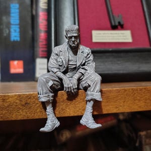 May include: A gray figurine of Frankenstein's monster sitting on a wooden surface. The figure is detailed with wrinkles and textured clothing. The monster is wearing a jacket, shirt, and boots. The background includes books and a framed key.