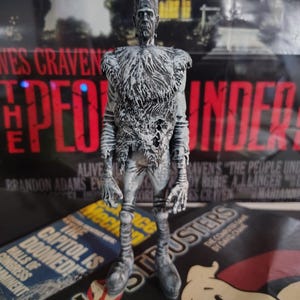 Puede incluir: Figura de acción de Frankenstein en gris y blanco. La figura tiene una apariencia texturizada con detalles visibles. El fondo incluye un cartel de película con el texto "The People Under the Stairs" y "Ghostbusters".