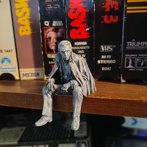 May include: A silver-colored figurine of a seated, elderly man with a cape, likely a character from a horror film. The figure is perched on a wooden shelf in front of a collection of VHS tapes, including titles like "Basket Case" and "Case 3."