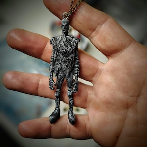 May include: A silver-colored pendant necklace featuring a detailed miniature figure of Frankenstein's monster. The figure has a textured fur-like chest and is suspended from a delicate chain. The pendant is held in a hand, showcasing its intricate design.