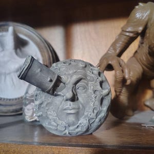 May include: A gray, spherical sculpture of a face with a beard and a telescope-like object protruding from it. The sculpture has a textured surface with craters and other details. The sculpture is on a wooden shelf.