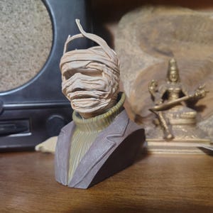 May include: A bust of a mummy figure with wrapped bandages around the head and face. The figure wears a green turtleneck sweater and a gray blazer. The bust is on a wooden surface.